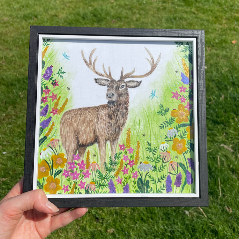 A hand holding a framed illustration of a stag among colourful flowers and greenery.