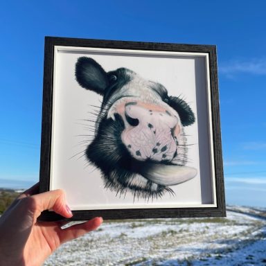 A person holds a framed illustration of a cow's face against a blue sky.