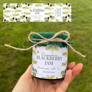 A hand holding a jar of blackberry jam with a green lid and decorative label.