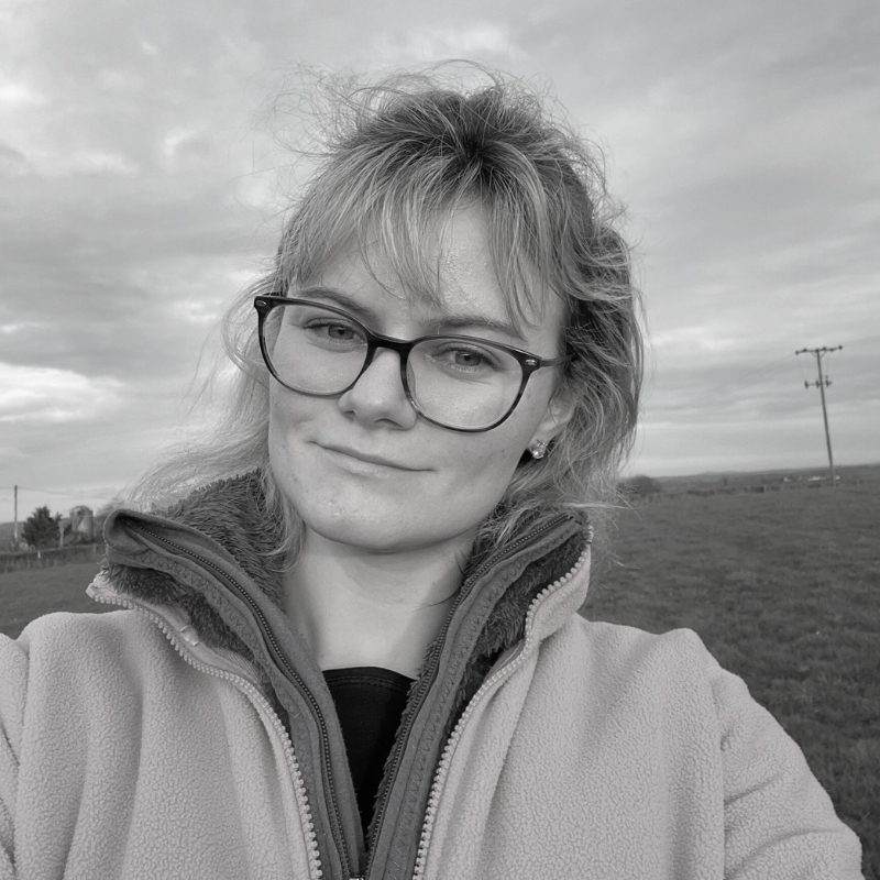 Chantelle with glasses and a light jacket smiles outdoors against a cloudy sky.