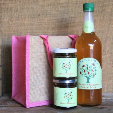 A gift set featuring a bottle of drink and two jars in a jute bag with pink handles.