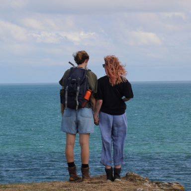 Two people holding hands, overlooking a calm sea from a rocky shoreline.