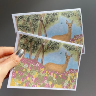 A person holding two illustrated cards featuring a deer in a floral landscape.