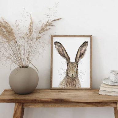 A framed artwork of a rabbit beside a vase and a cup on a wooden table.