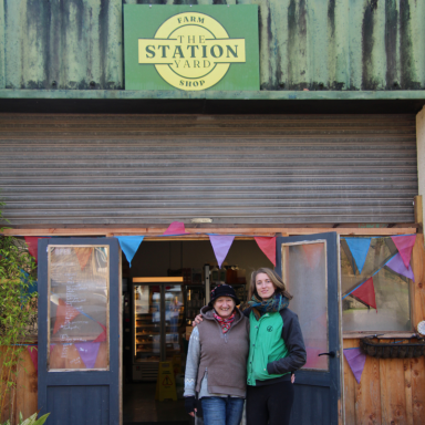 Two women stand outside a rustic shop entrance, decorated with colourful bunting.