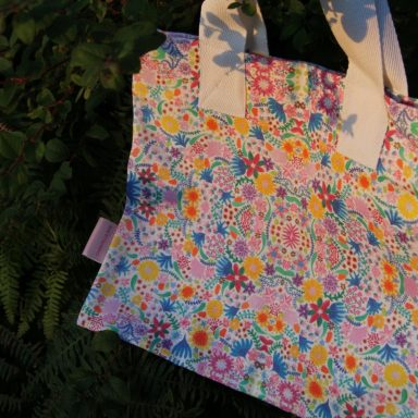 Colourful floral-patterned tote bag with white handles, resting on greenery.