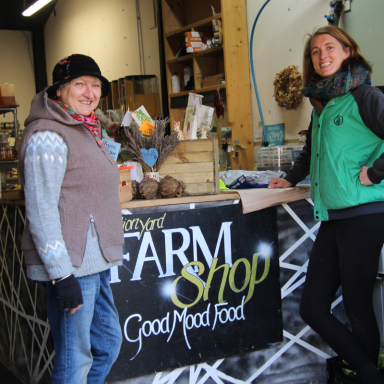 Two women pose in a farm shop, smiling beside a counter with fresh produce and goods.