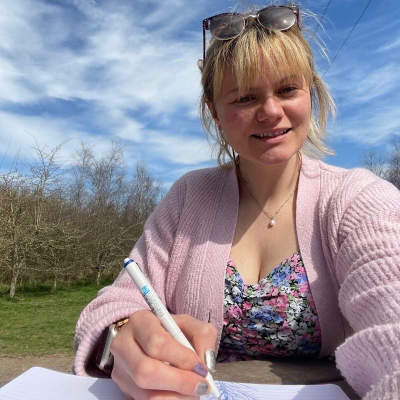 Chantelle drawing on paper outdoors, dressed colourfully with a natural backdrop and bright sky.